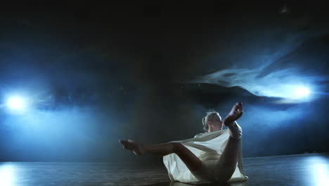 Ein-Modernes-Tanzmädchen-In-Einem-Weißen-Kleid-Tanzt-Ein-Modernes-Ballett-Und-Springt-Auf-Der-Bühne-Mit-Rauch-Im-Blauen-Scheinwerferlicht.
